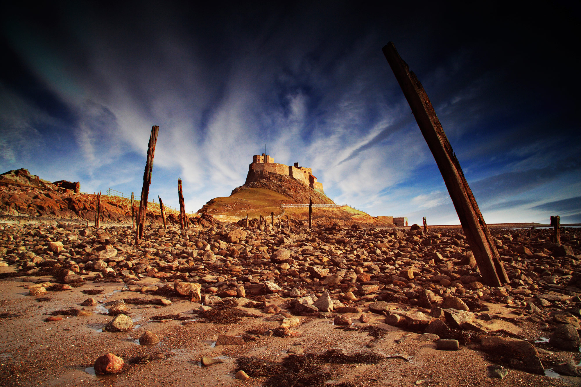 Lindisfarne Holy Island - rocky beach with the ancient wooden posts in the foreground leading the eye to the magnificent castle in the background.